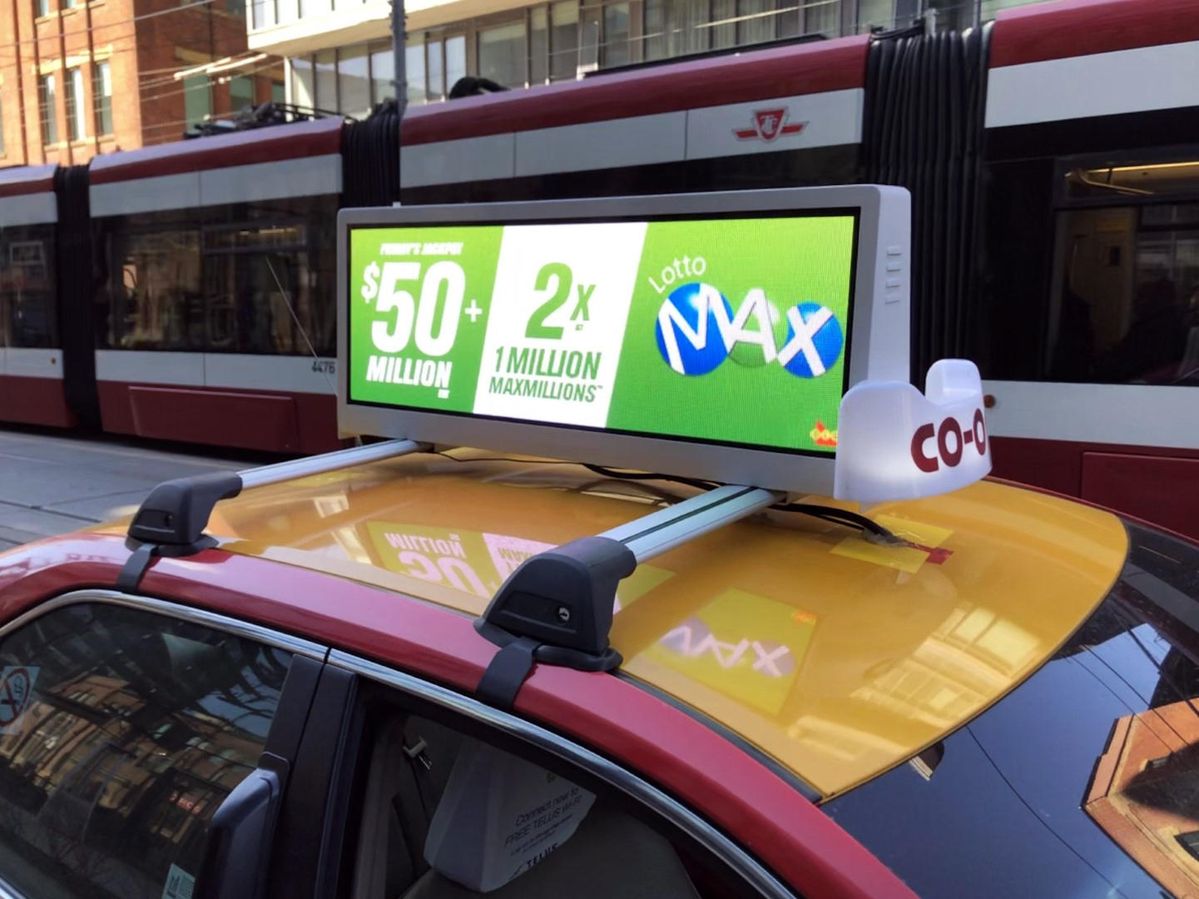 A P2 LED screen mounted on a taxi roof, displaying a vibrant lottery advertisement with high clarity, seen with a streetcar in the background.